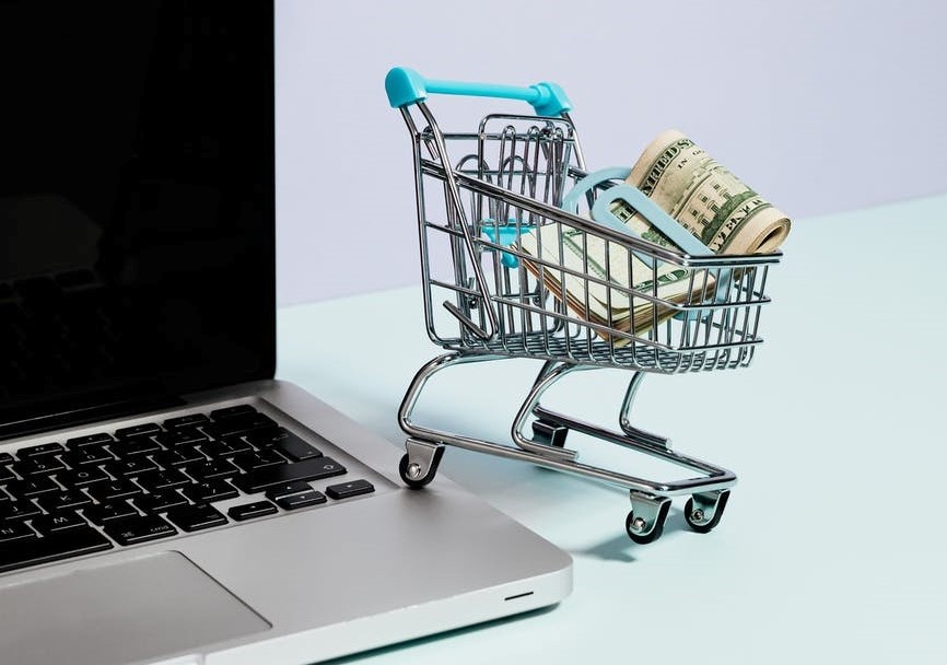 carrito de supermercado miniatura con fajo de billetes junto a laptop